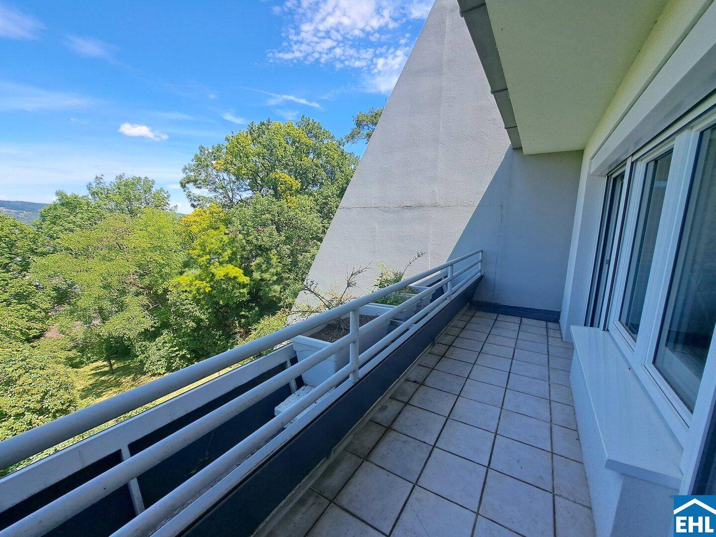 Balkon mit Fliesenboden und weitem Blick ins Grüne unter blauem Himmel.