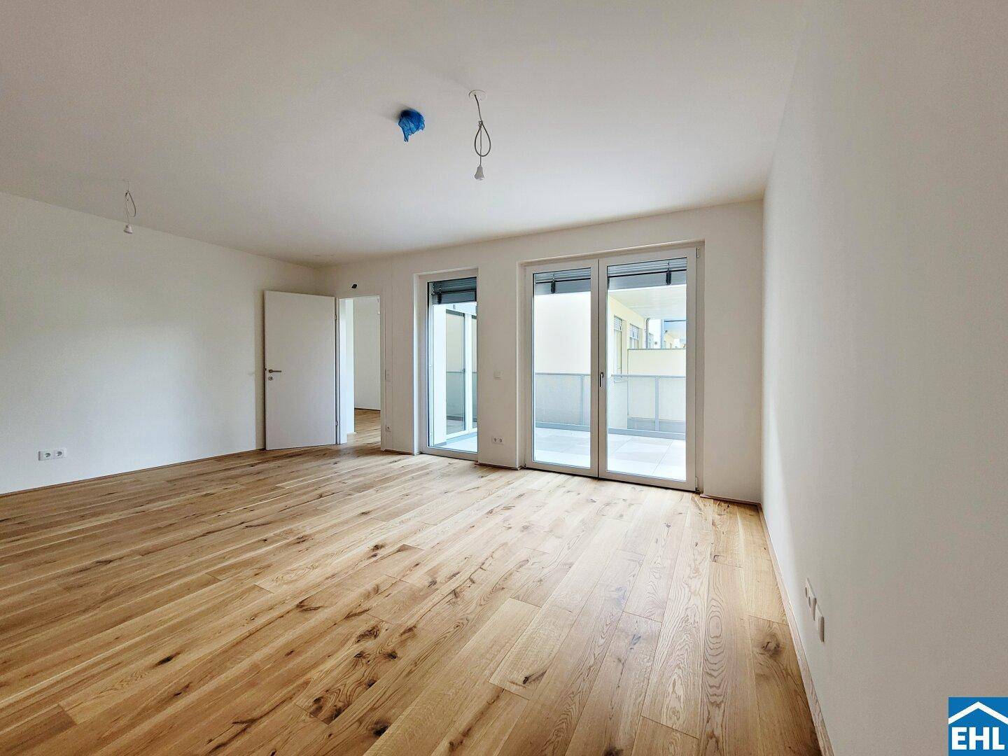 Geräumiges Wohnzimmer mit Holzboden und großen Fenstertüren, die auf einen Balkon führen.