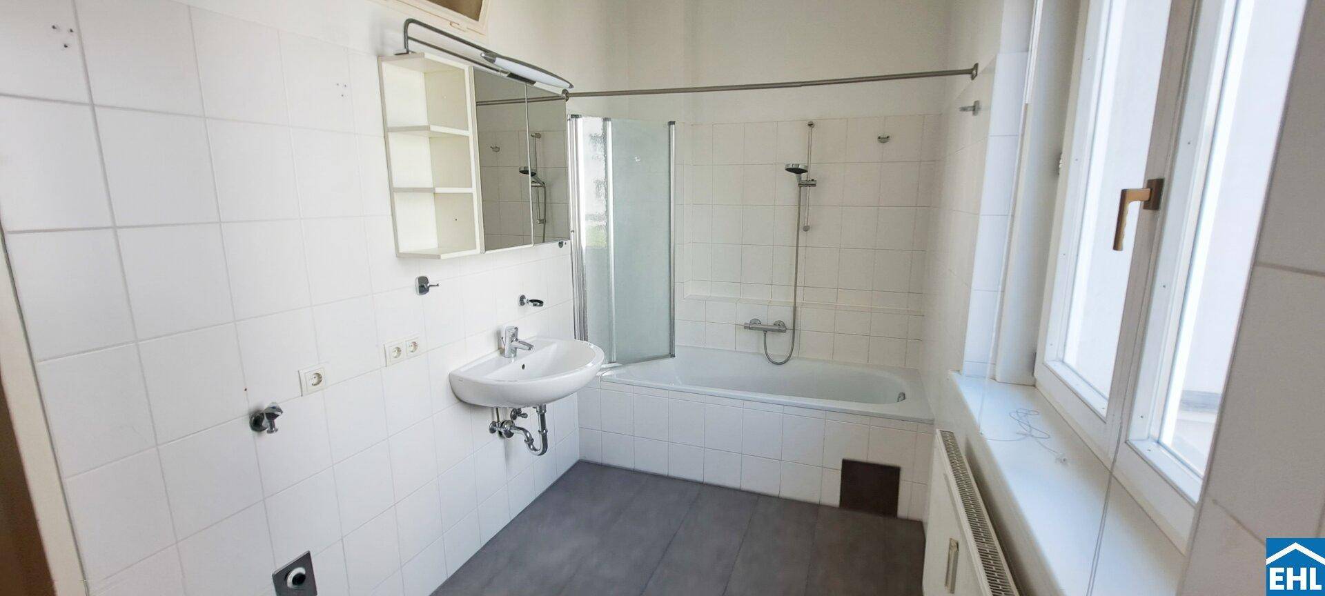 Badezimmer mit Badewanne, Waschbecken, Spiegelschrank und Fenster, hell gefliest.