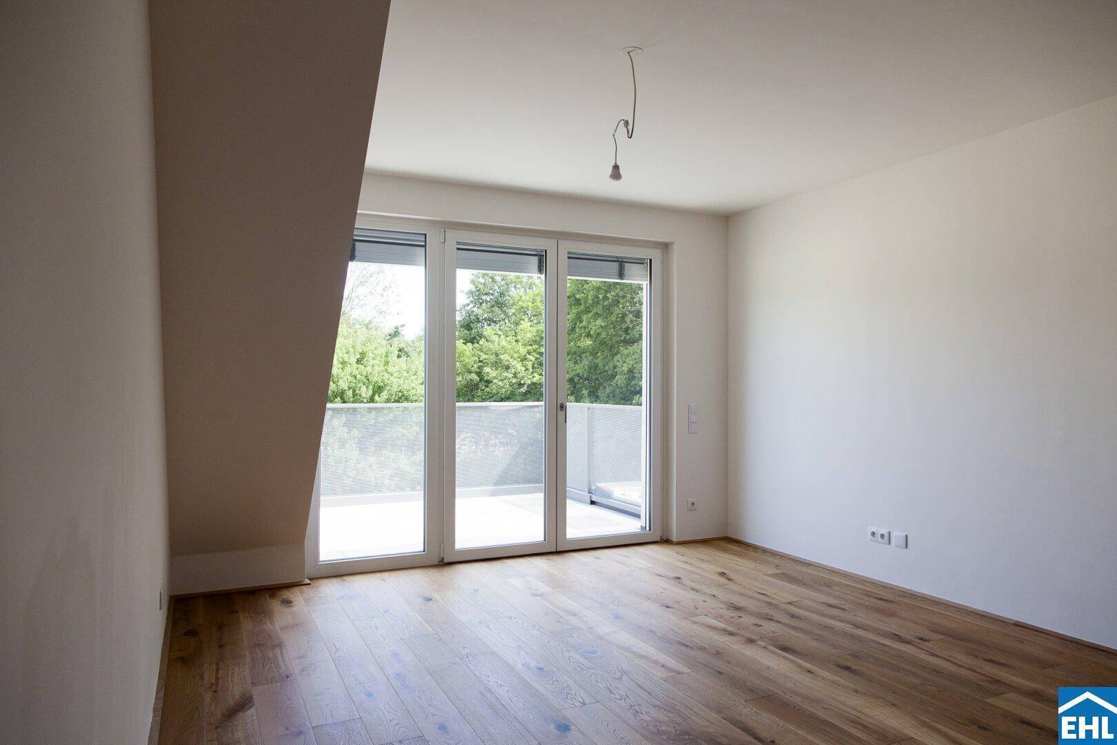 Geräumiger, heller Raum mit Holzboden und großen Balkontüren mit Blick ins Grüne.