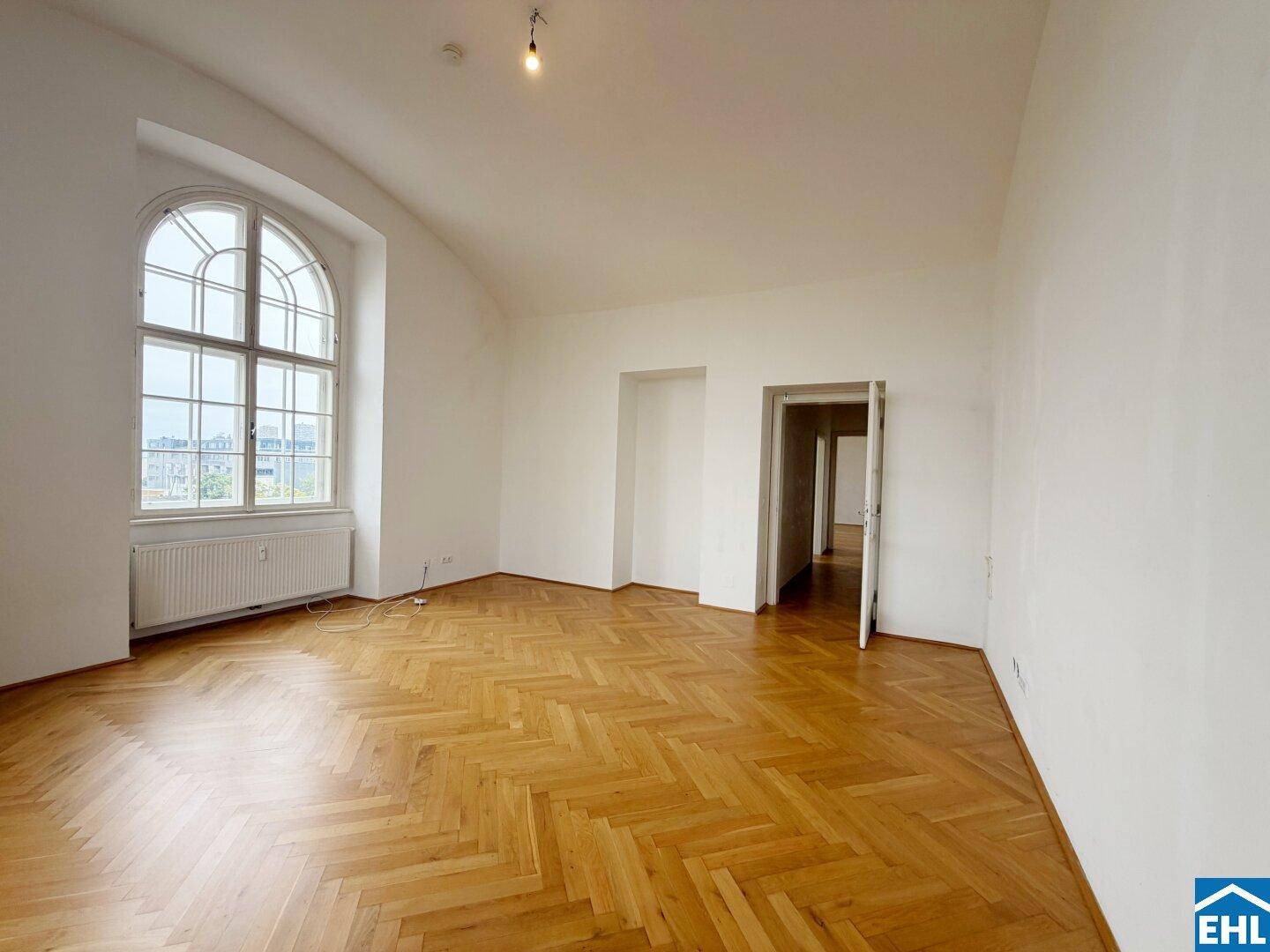 Geräumiges Zimmer mit Fischgrätparkett, hohen Decken und Zugang zu weiteren Räumen.