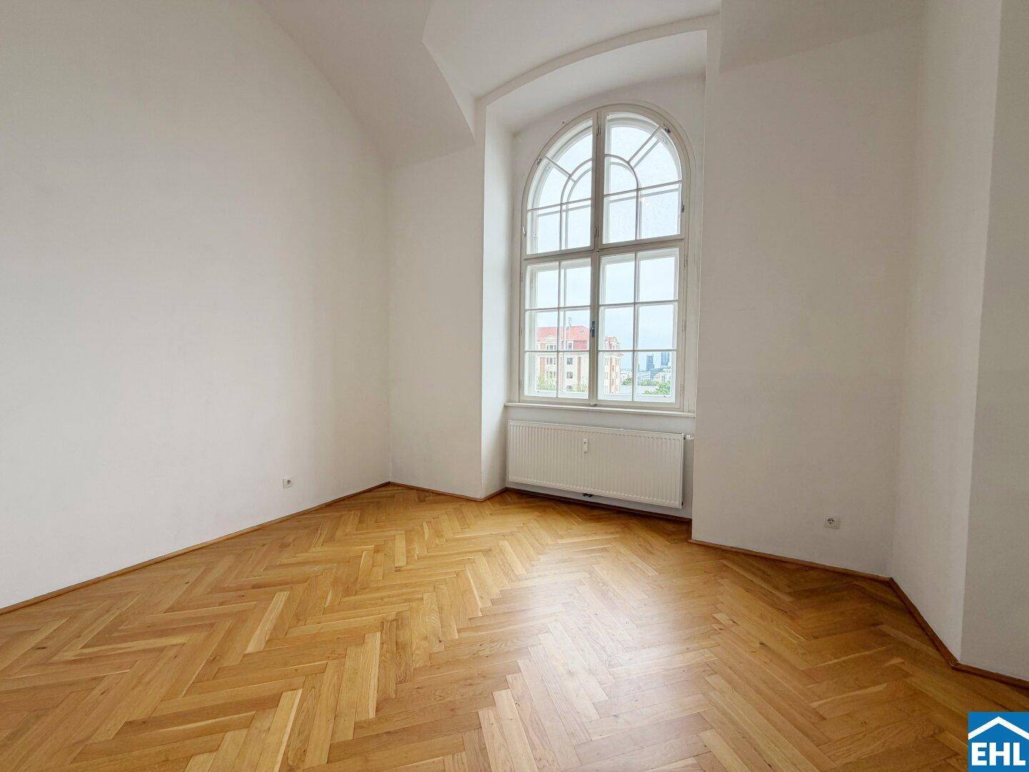 Lichtdurchflutetes Zimmer mit klassischem Fischgrätparkett und einem großen Rundbogenfenster.