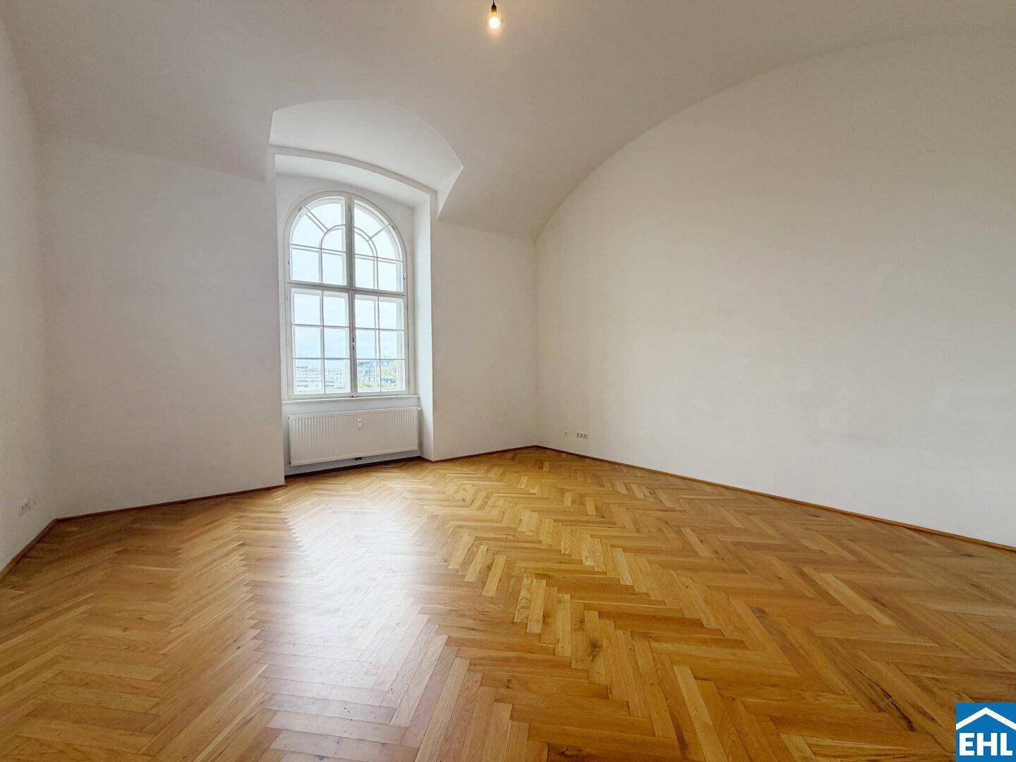 Heller Raum mit Fischgrätparkett und einem markanten Rundbogenfenster, das viel Licht spendet.