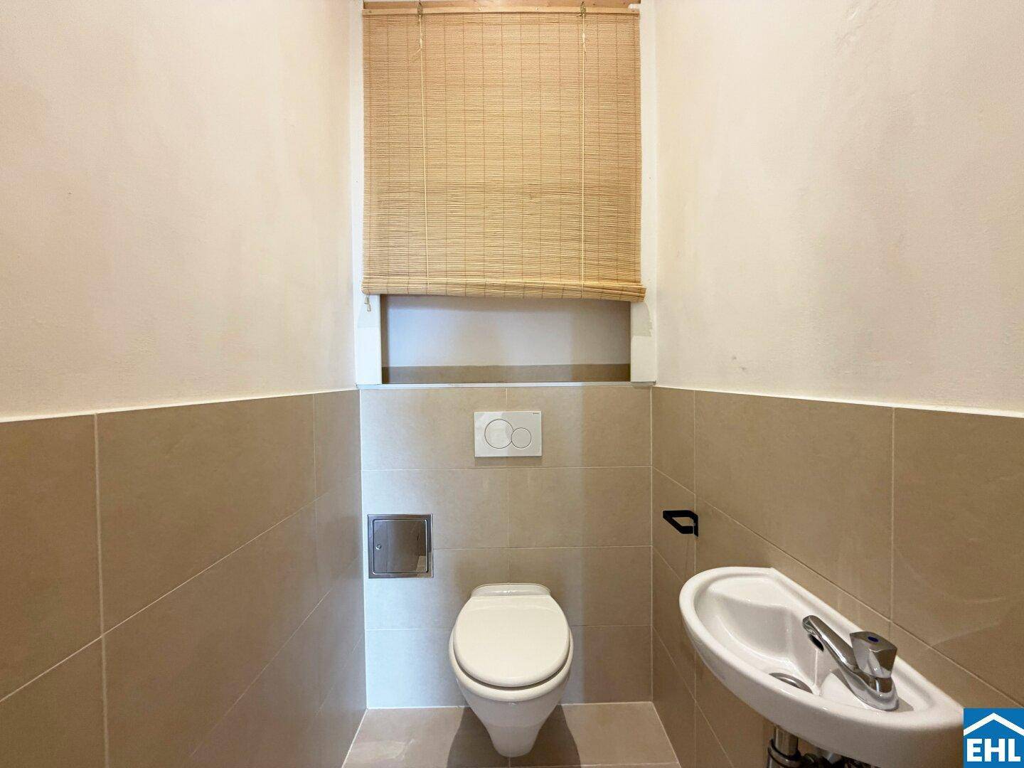 Separates WC mit wandhängender Toilette, kleinem Waschbecken und Fenster mit Jalousie.