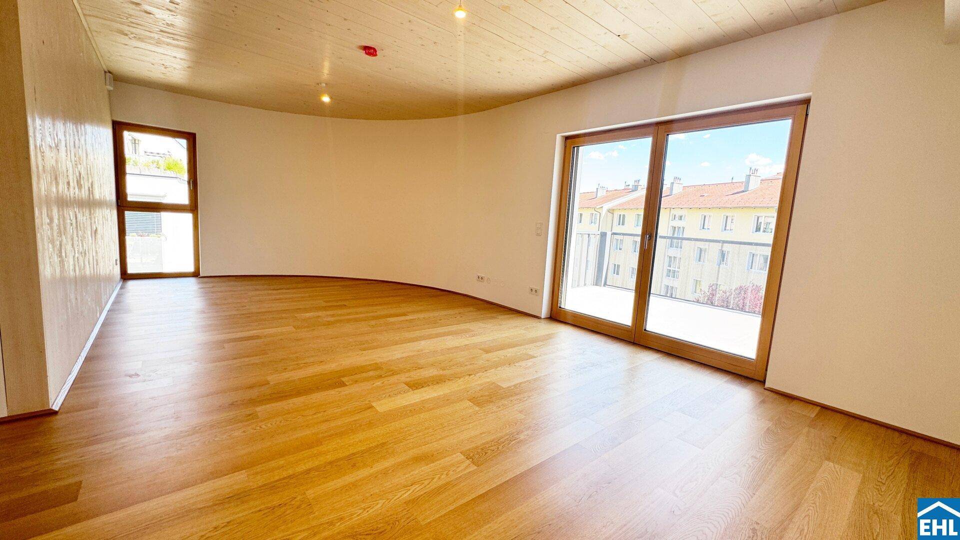 Lichtdurchflutetes Wohnzimmer mit Holzboden und weitem Blick auf die umliegenden Gebäude und die Natur.