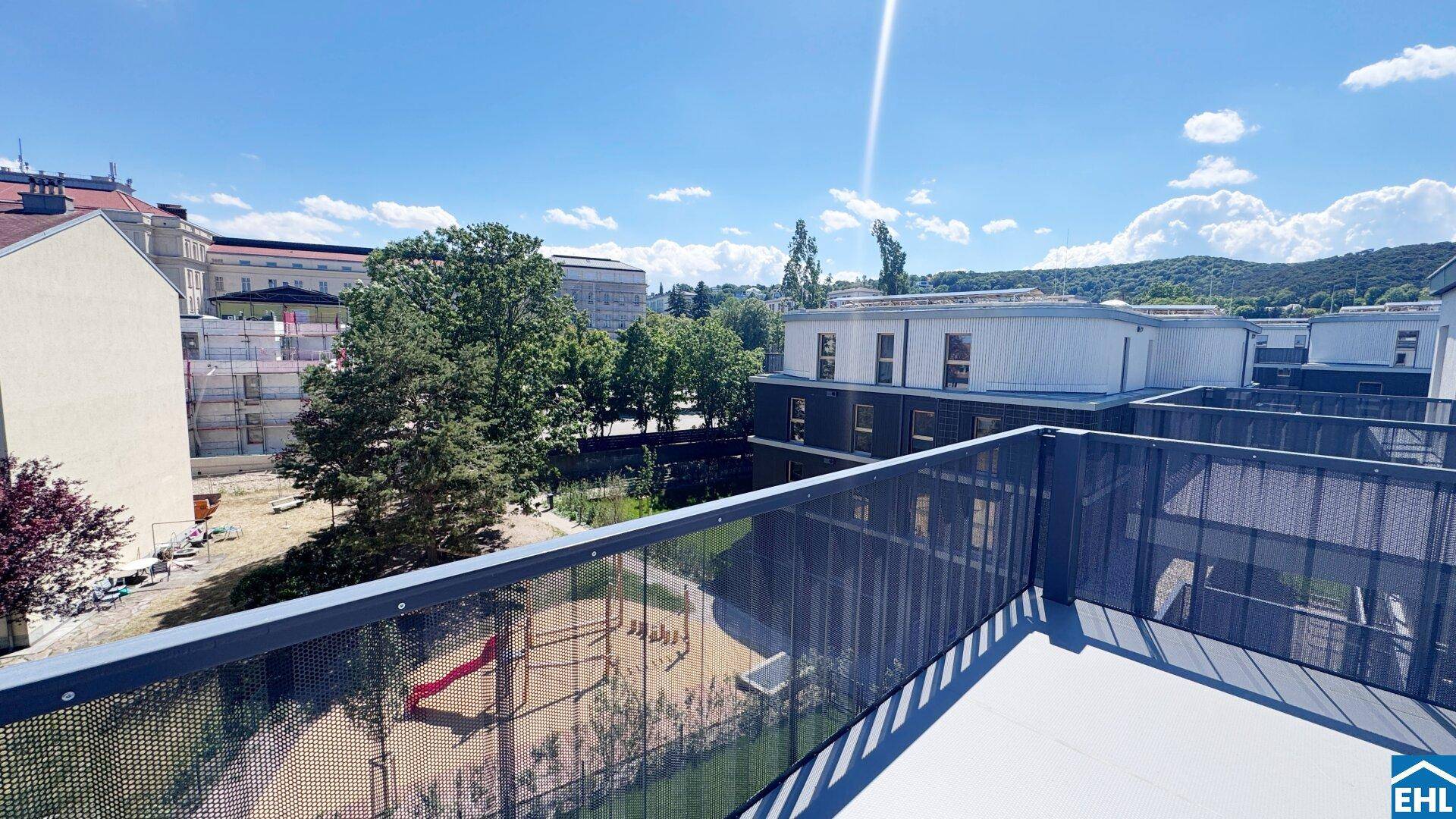 Großer Balkon mit Blick auf die umliegenden Gebäude, Bäume und einen Spielplatz unter blauem Himmel.