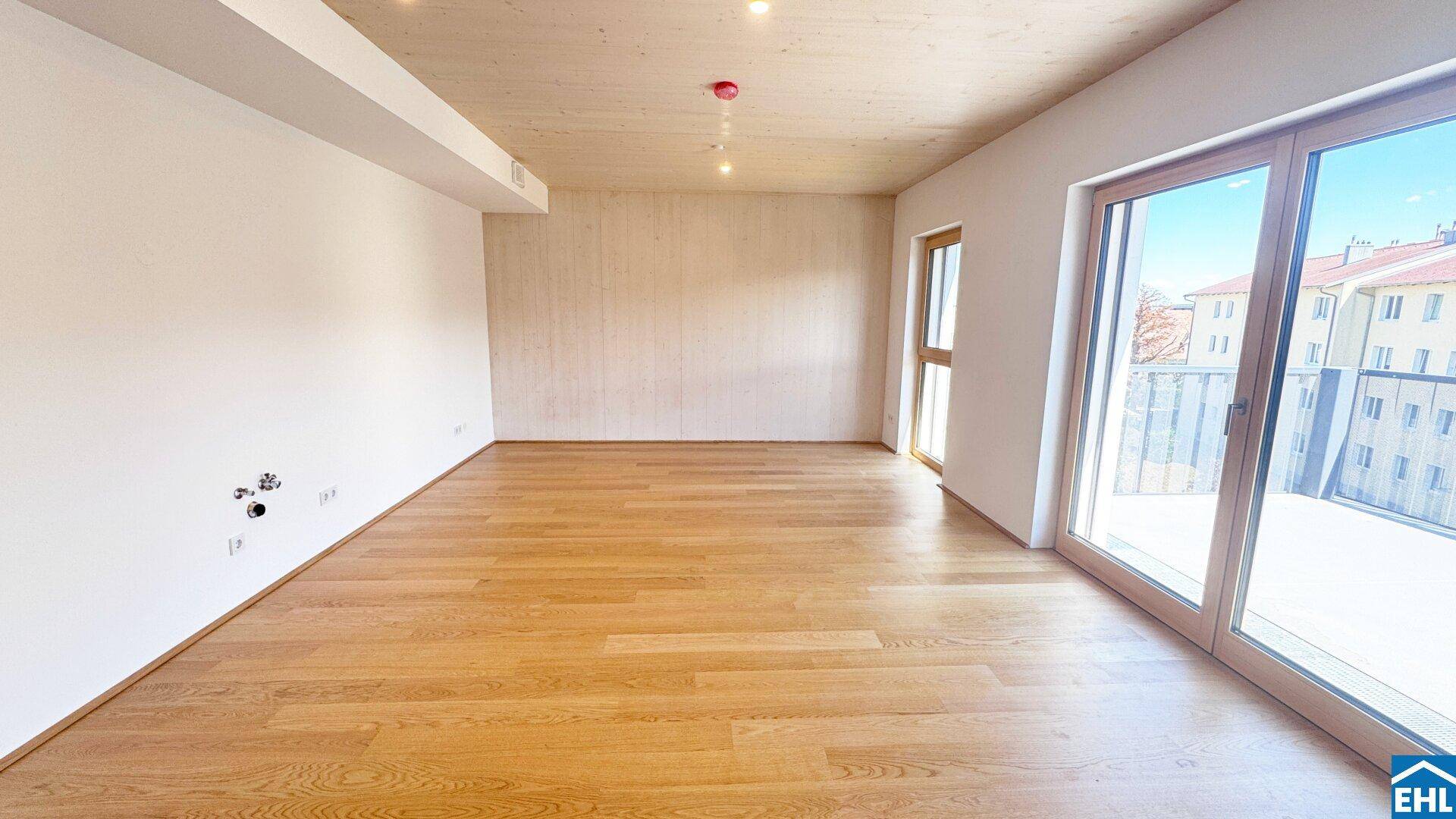 Geräumiger, unmöblierter Raum mit Holzboden, Holzdecke und großen Fenstern zum Balkon.
