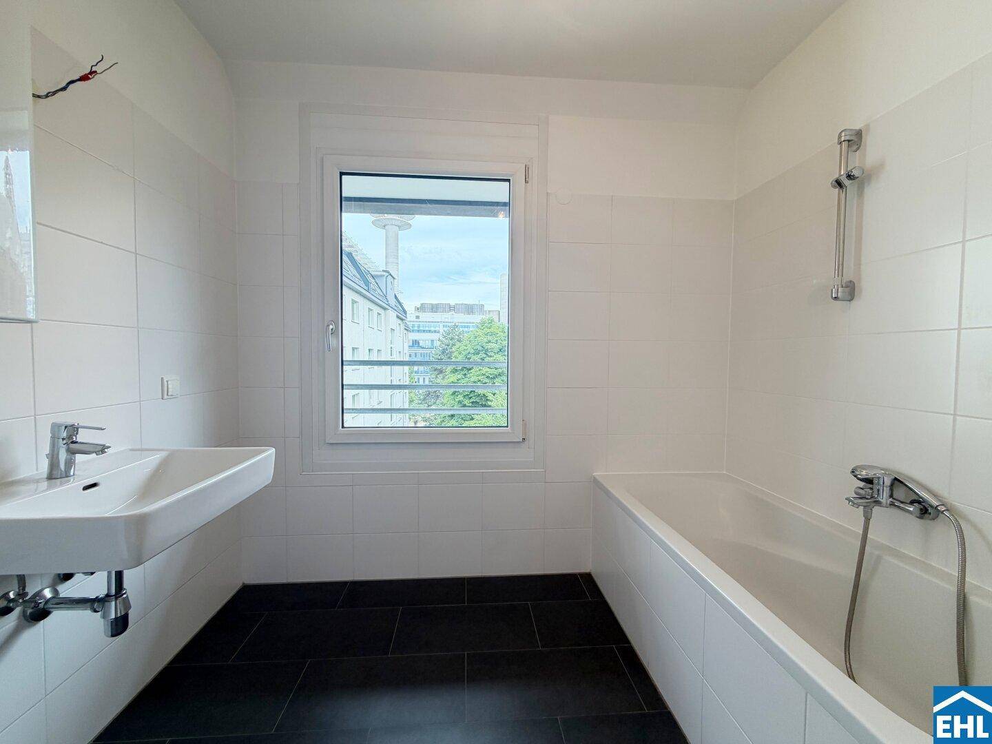 Modernes Badezimmer mit weißen Fliesen, dunklem Boden, Badewanne und Fenster für Tageslicht.