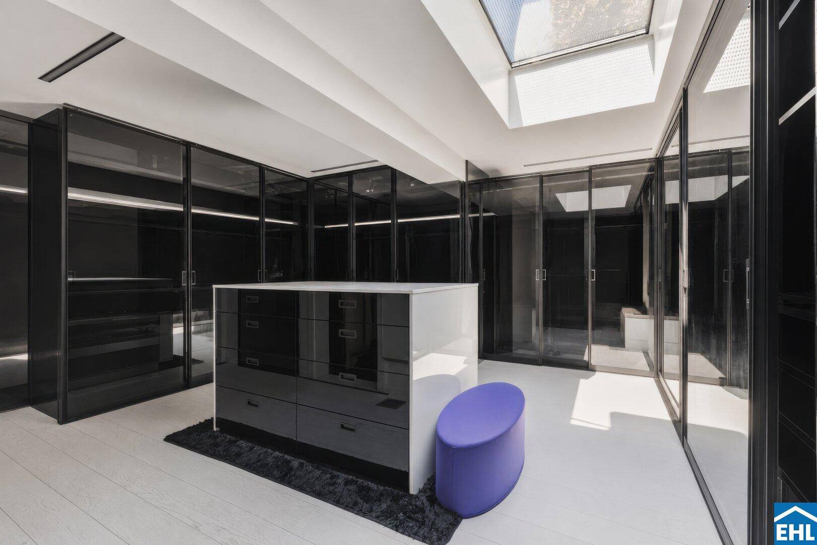 Geräumiger begehbarer Kleiderschrank mit schwarzen Glasfronten, Mittelinsel und Oberlicht.