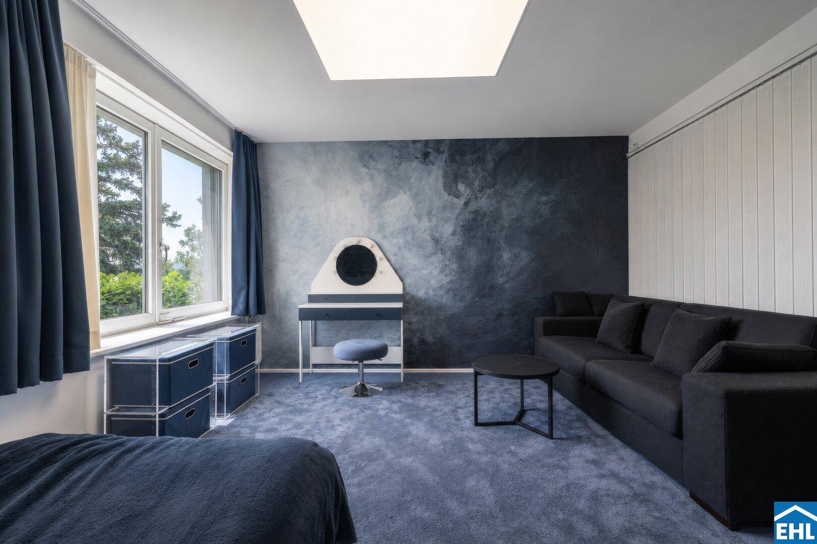 Modernes Schlafzimmer mit blauer Akzentwand, bequemer Couch und Blick ins Grüne.