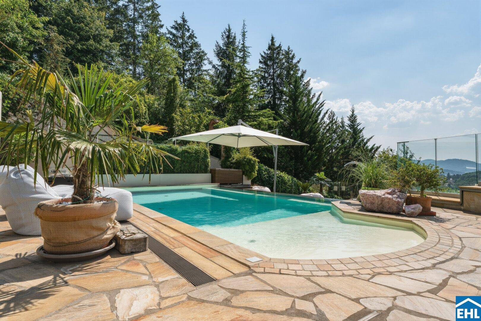 Großer Swimmingpool mit Liegeflächen, Palmen und Sonnenschirm, umgeben von Natursteinplatten.