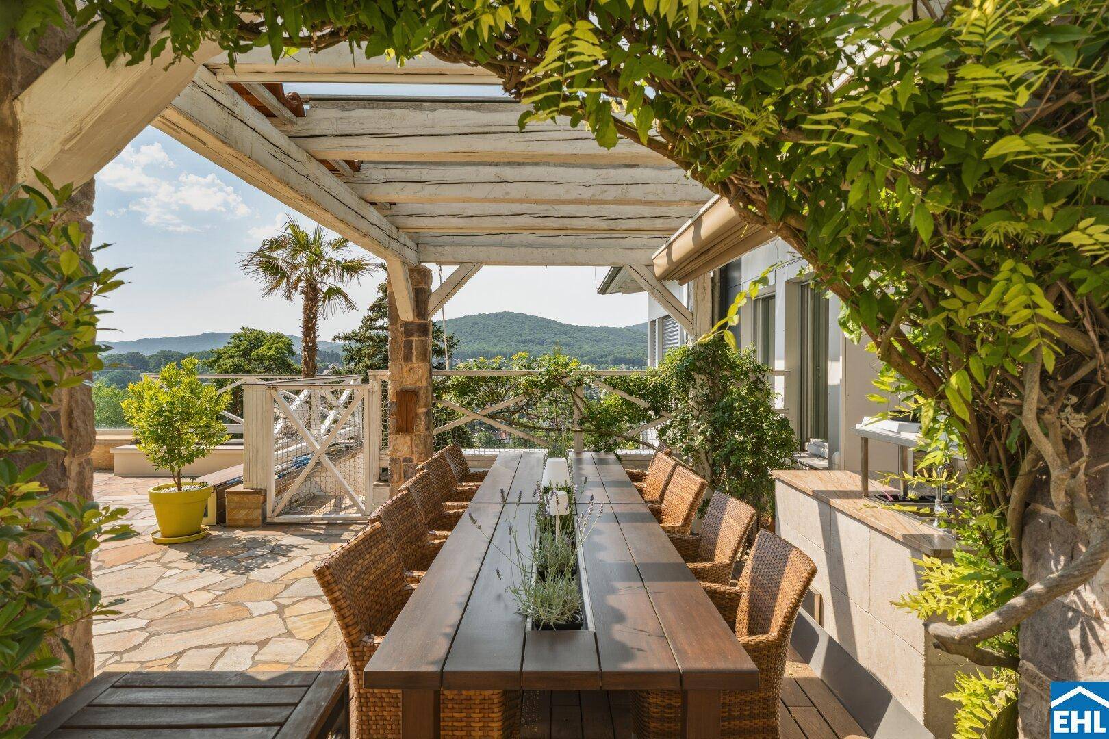 Stilvoller Essbereich im Freien mit großem Holztisch und Rattanmöbeln unter einer bewachsenen Pergola.