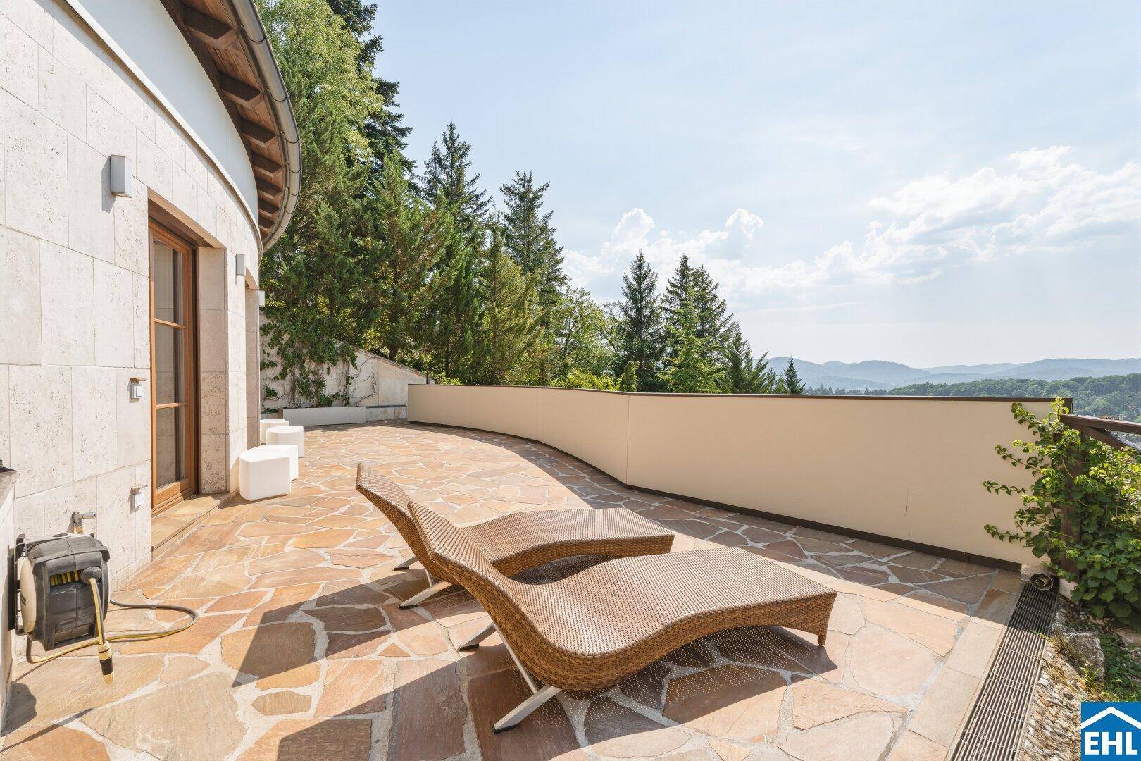 Weitläufige, geschwungene Terrasse mit Natursteinboden und Sonnenliegen, umgeben von viel Grün.