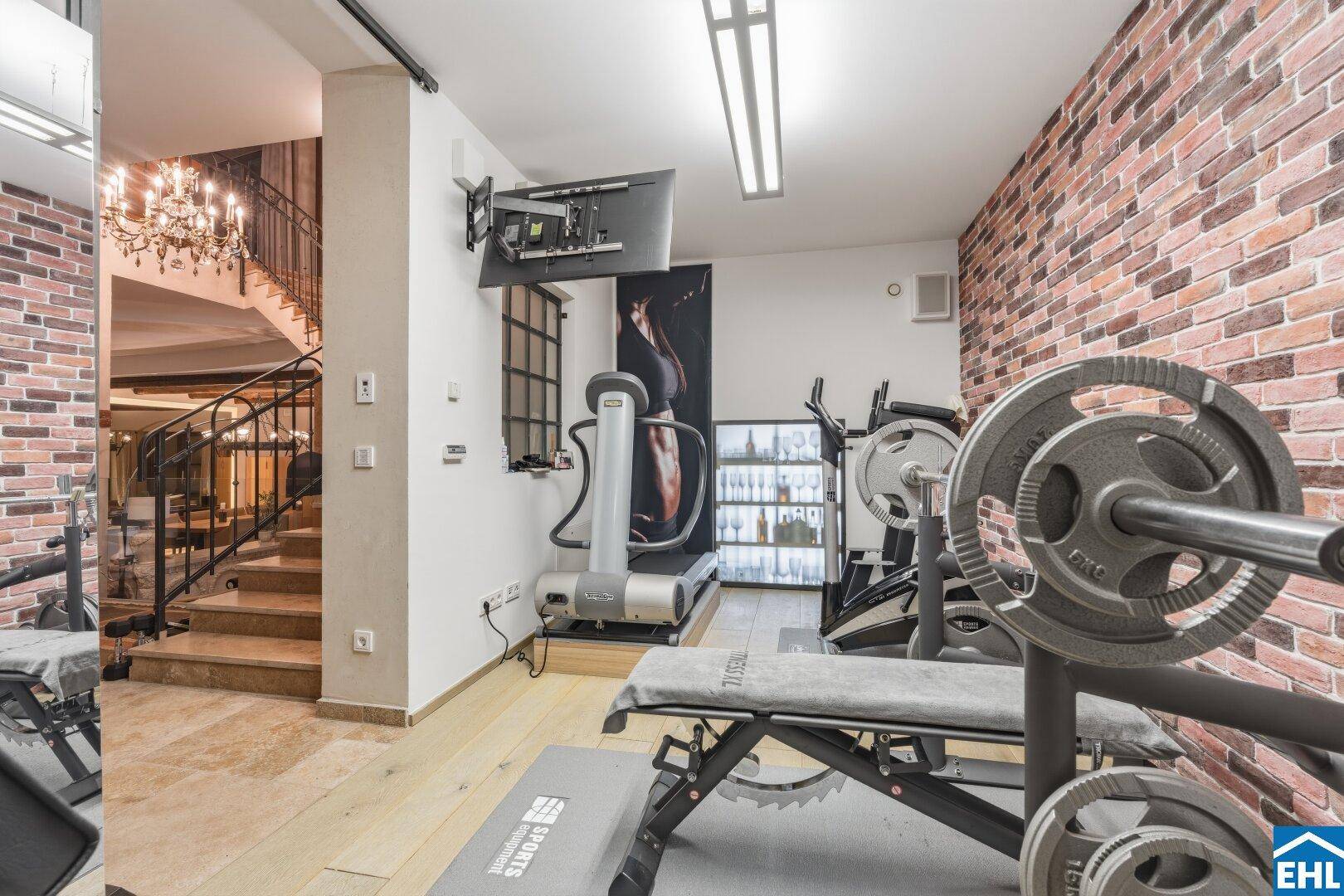 Gut ausgestatteter Fitnessraum mit Trainingsgeräten, Ziegelwand und Blick auf eine elegante Treppe.