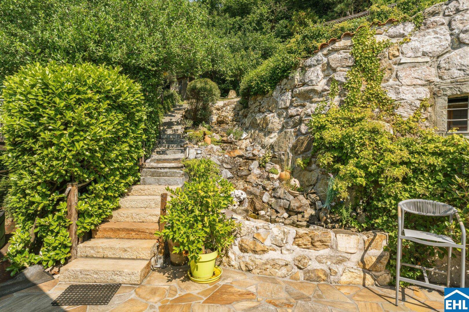 Steintreppe führt durch den terrassierten Garten mit Natursteinmauer und kleinem Wasserfall.