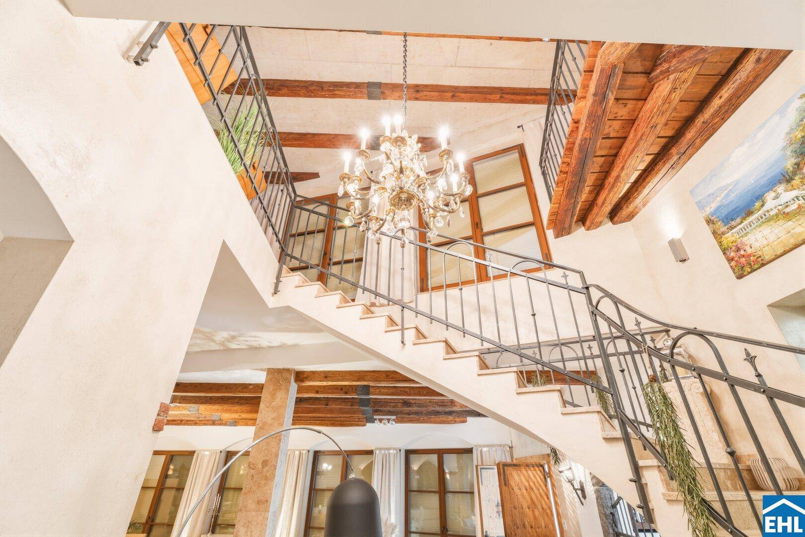 Elegantes Treppenhaus mit schmiedeeisernem Geländer, Kronleuchter und Holzbalken.