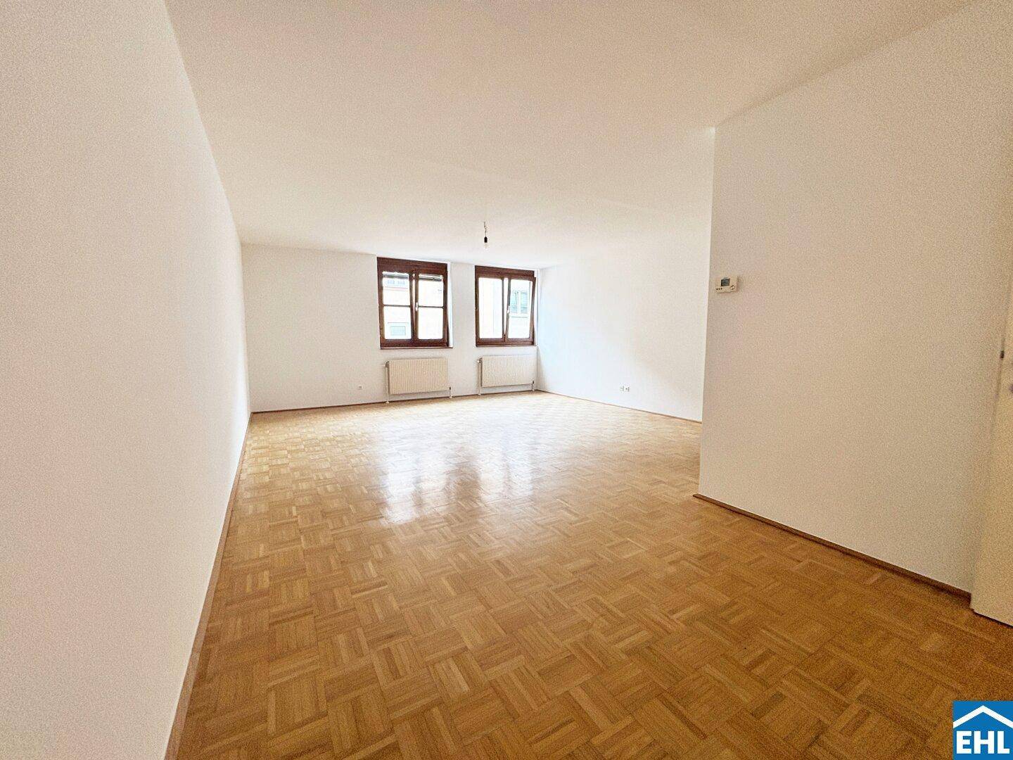 Geräumiger Wohnraum mit Fischgrätparkett und großen Fenstern, die viel Licht hereinlassen.