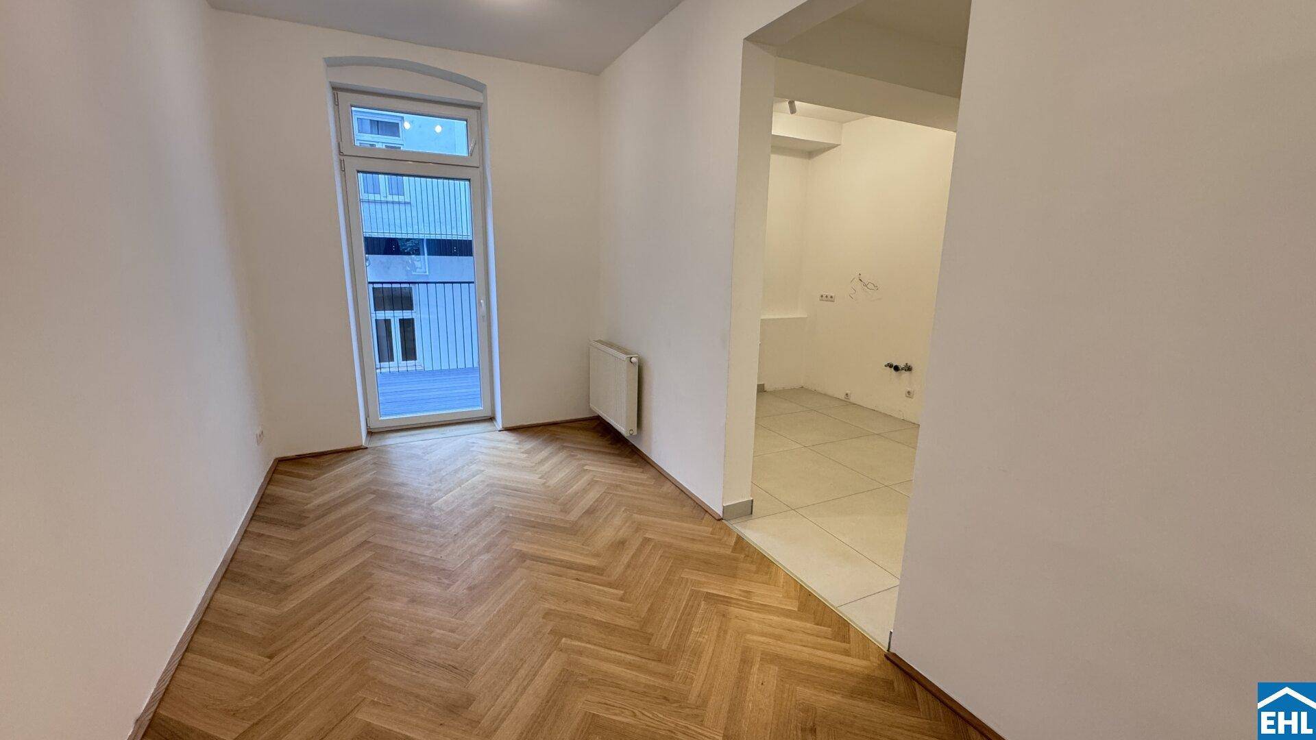 Helles Zimmer mit Fischgrätparkettboden und Zugang zu einem kleinen Balkon, angrenzend an einen gefliesten Bereich.