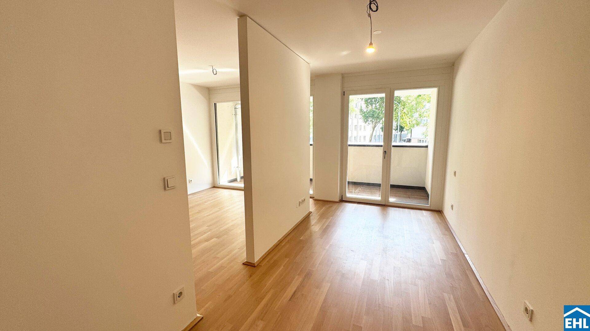 Leerer Wohnraum mit hellem Parkettboden und Zugang zu einem weiteren Zimmer und Balkon.