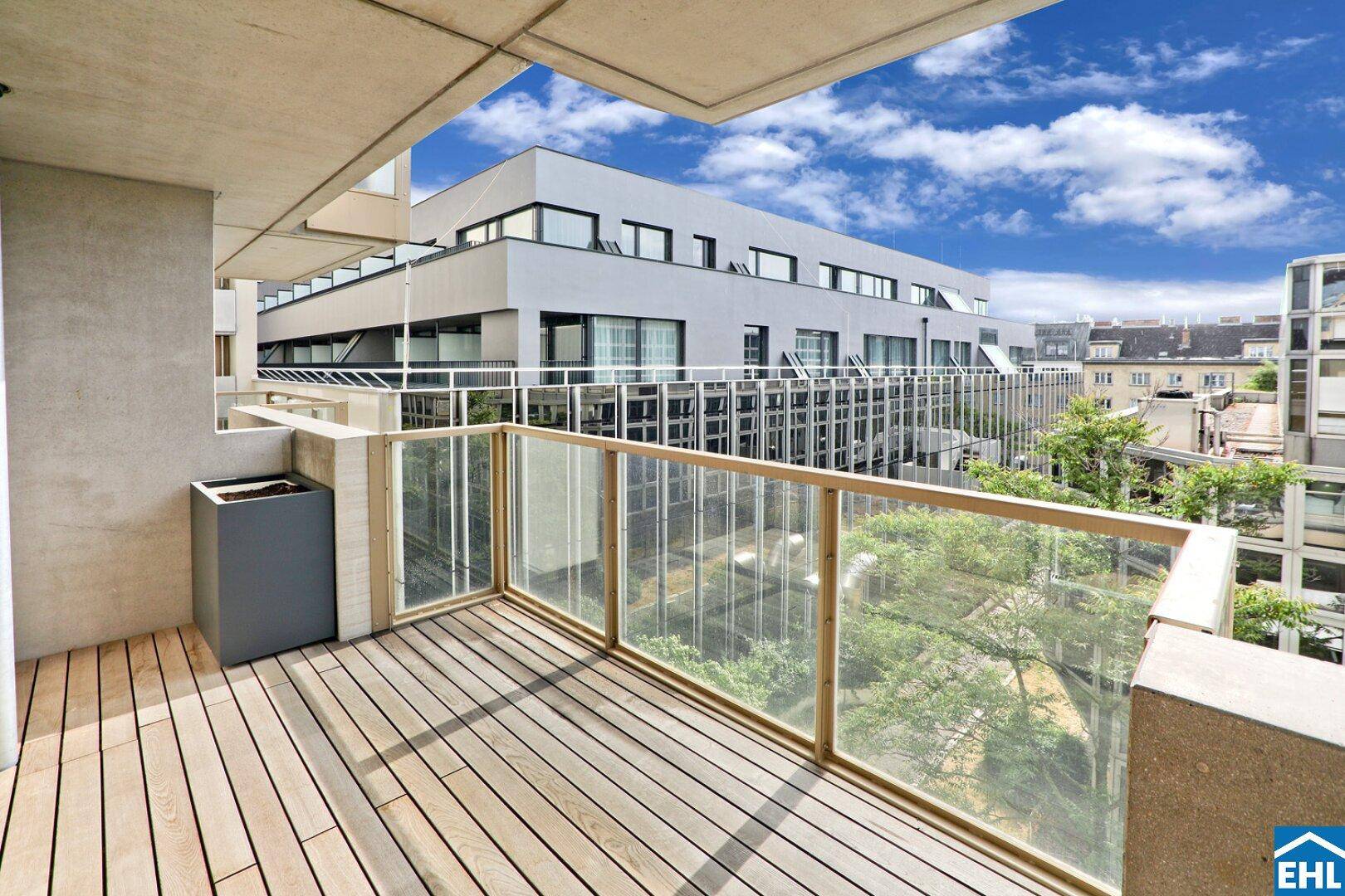 Sonniger Balkon mit Holzboden und Glasgeländer, umgeben von moderner Wohnbebauung.