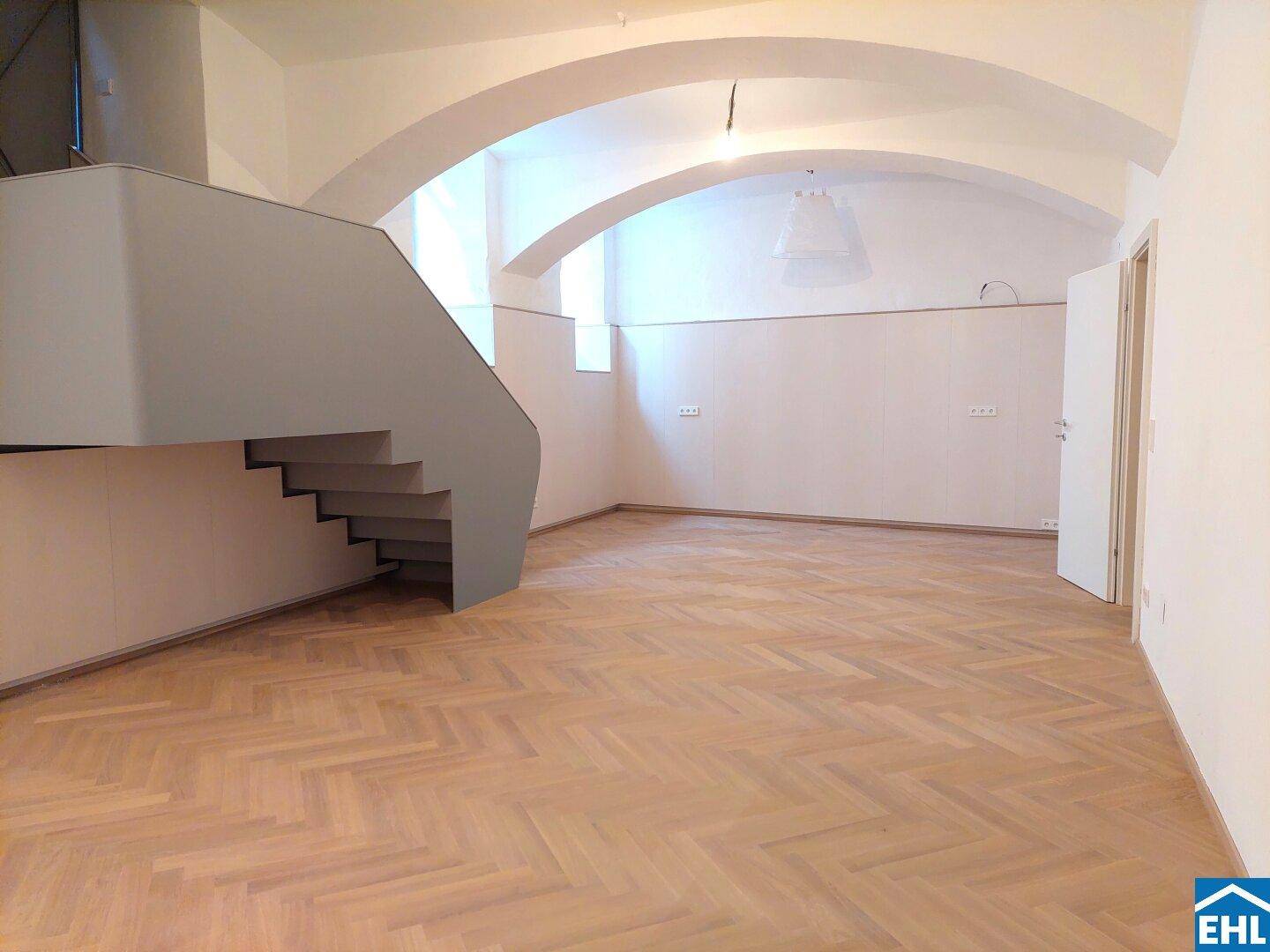 Heller Innenraum mit Fischgrätparkett, gewölbten Decken und einer modernen Treppe zu einer oberen Etage.