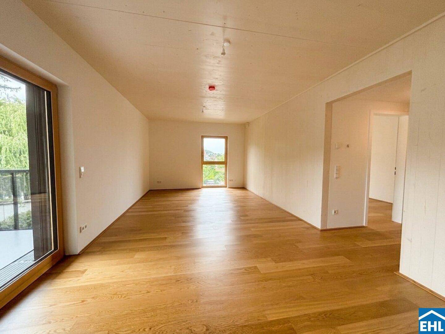 Geräumiger, heller Raum mit Holzboden und zwei Fenstern, eines davon mit Balkonzugang.