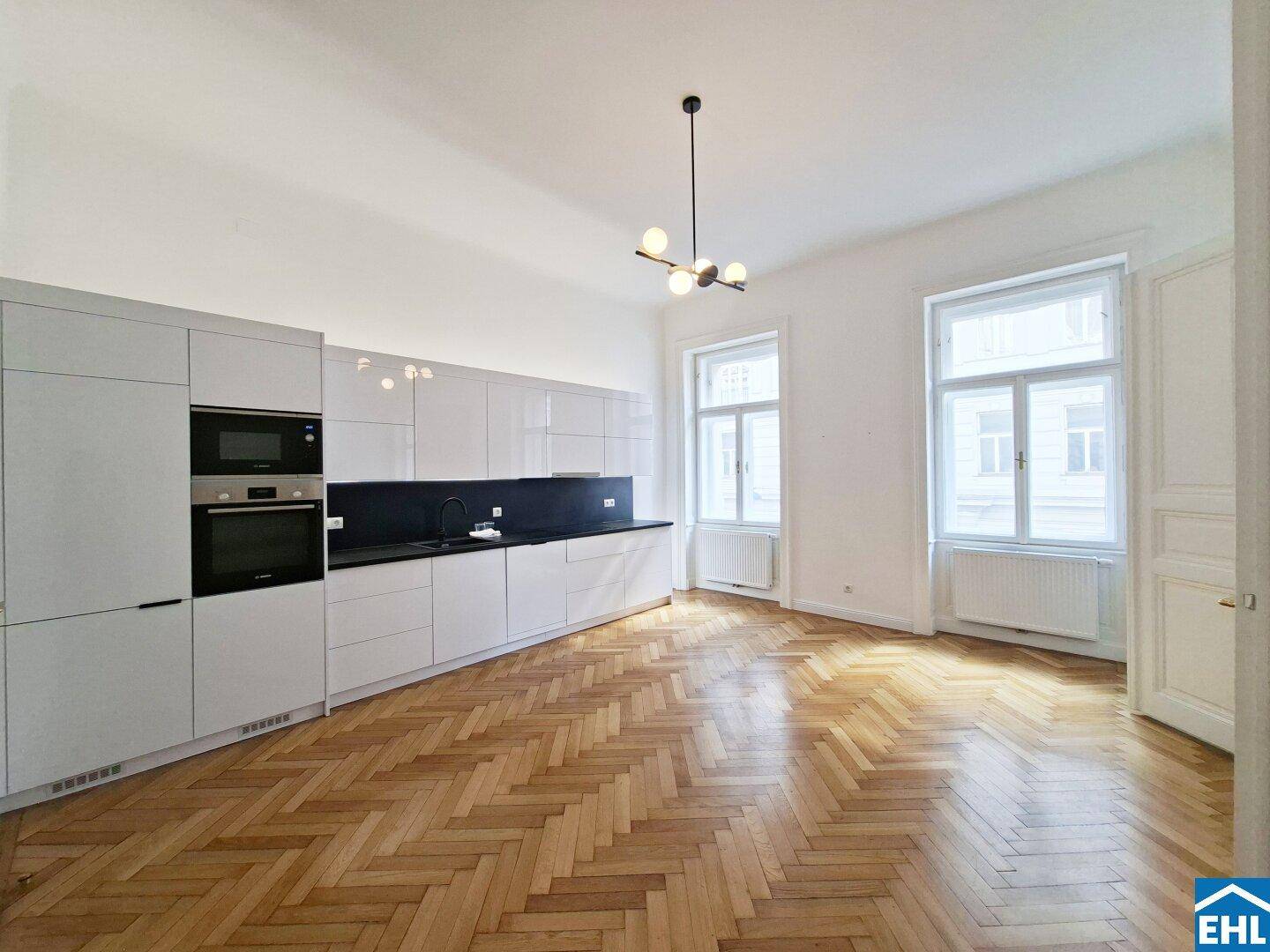 Geräumige Küche mit weißen Hochglanzfronten, schwarzer Arbeitsplatte und großen Fenstern.