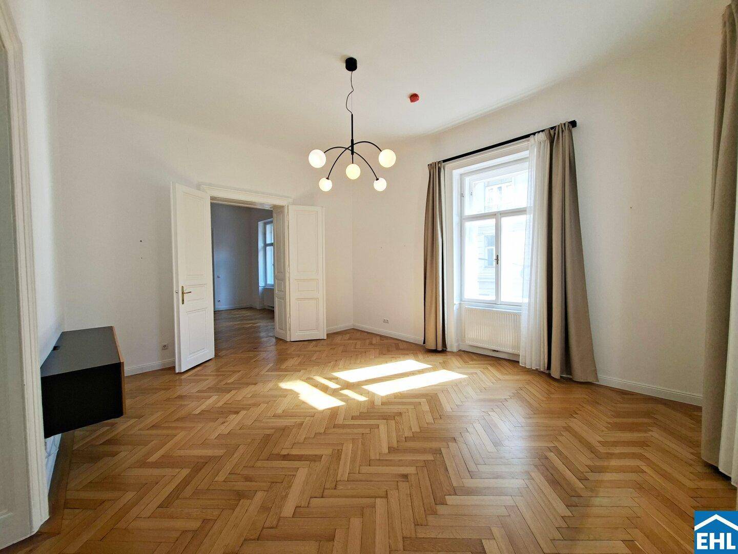 Geräumiges Wohnzimmer mit Fischgrätparkett, hohen Decken und viel natürlichem Licht.