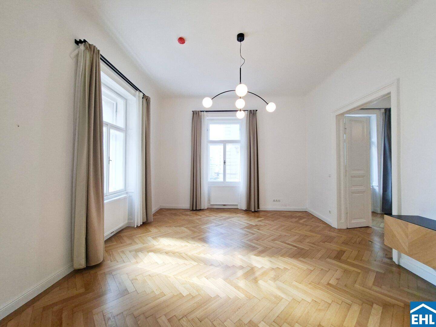 Lichtdurchflutetes Wohnzimmer mit elegantem Fischgrätparkett und hohen Fenstern mit Vorhängen.