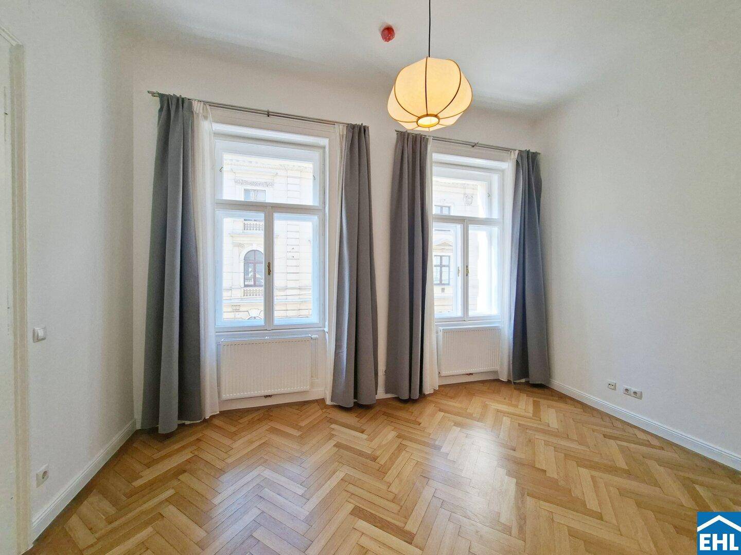 Helles Schlafzimmer mit Fischgrätparkett, hohen Fenstern und grauen Vorhängen.