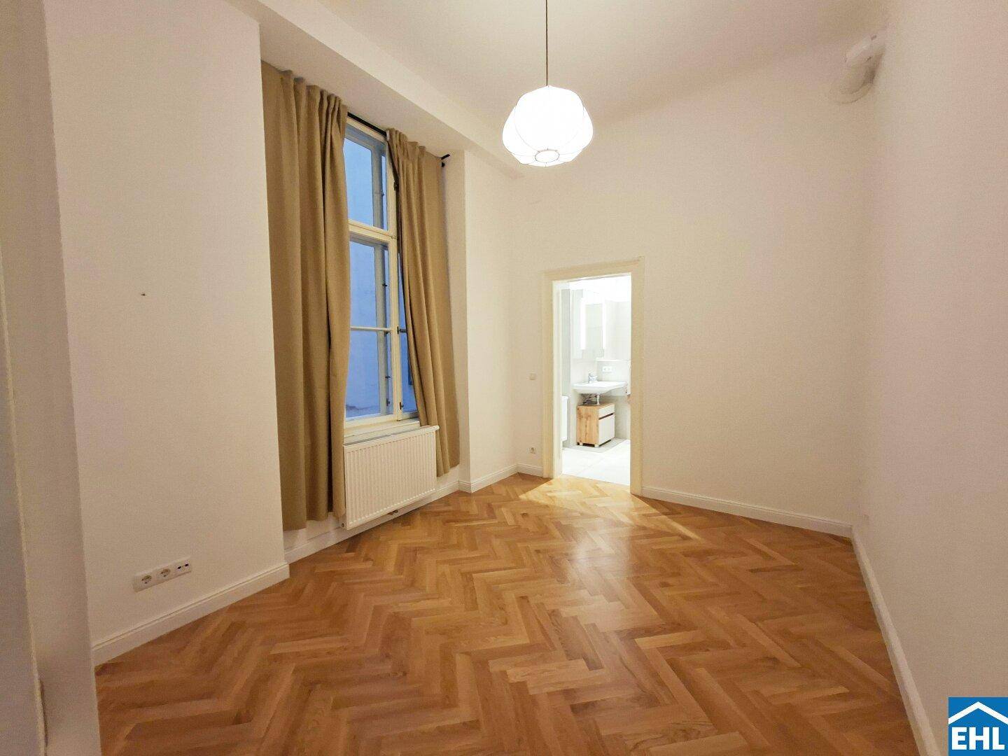 Helles Schlafzimmer mit Fischgrätparkett, hohen Decken und Zugang zu einem Badezimmer.