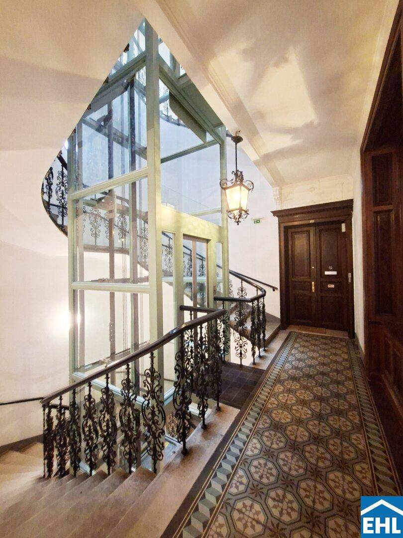 Historisches Treppenhaus mit elegantem Aufzug, kunstvollen Geländern und gemustertem Boden.