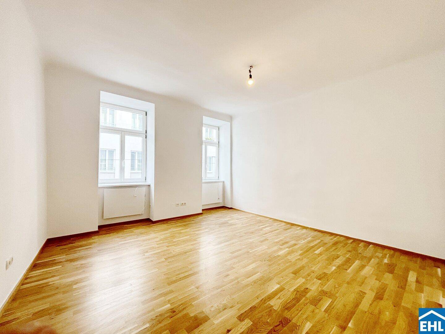 Heller, leerer Raum mit Holzboden und zwei großen Fenstern, ideal als Wohnzimmer.