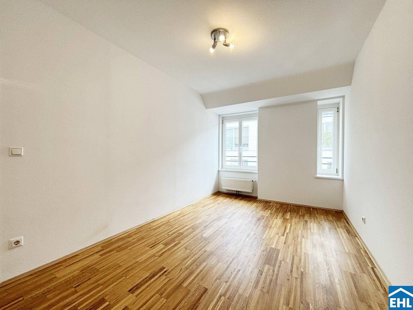 Helles Schlafzimmer mit Parkettboden, weißen Wänden und zwei Fenstern zur Straße.