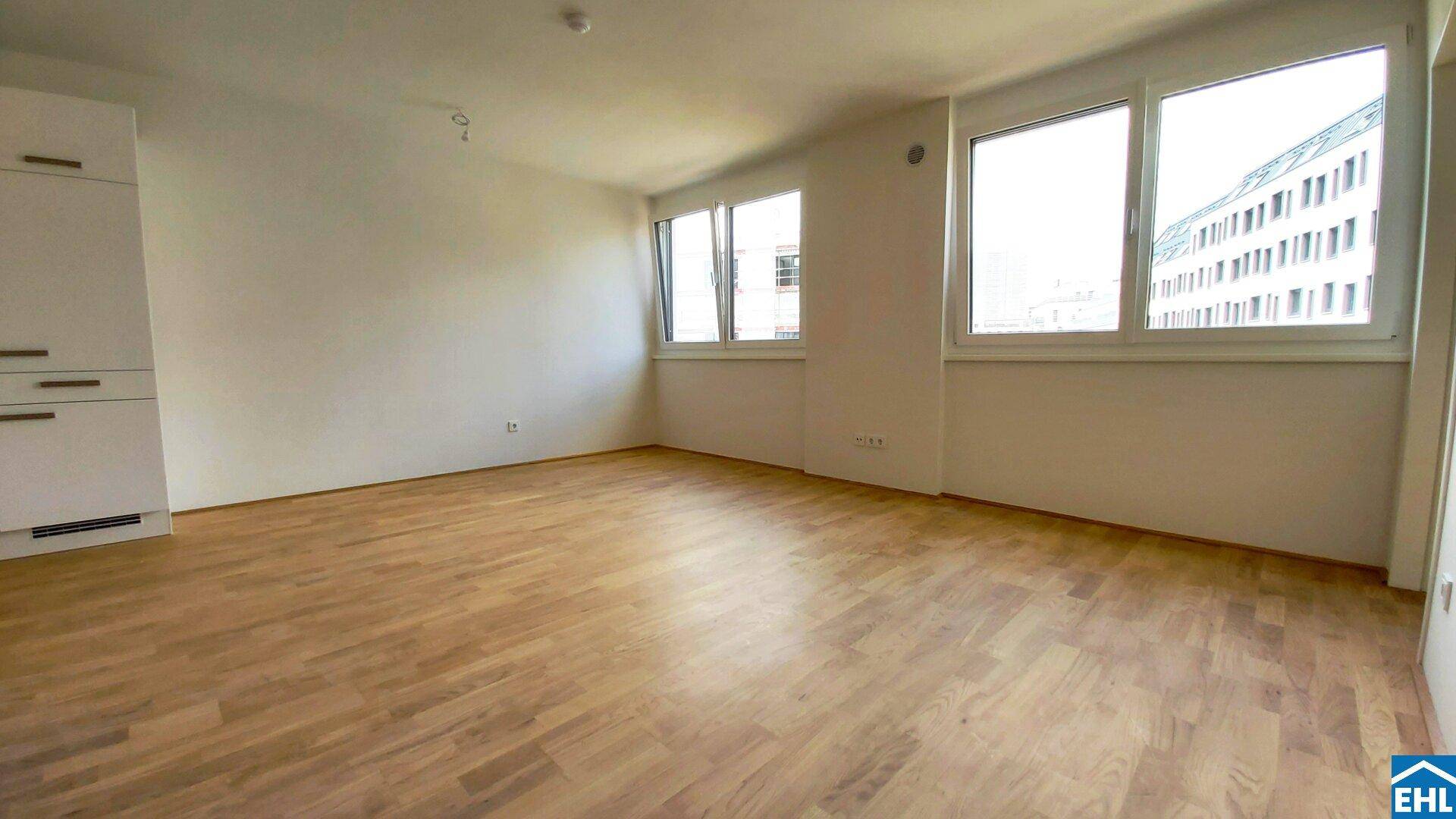 Leerer Raum mit großen Fenstern, hellem Holzfußboden und Blick auf ein Gebäude.