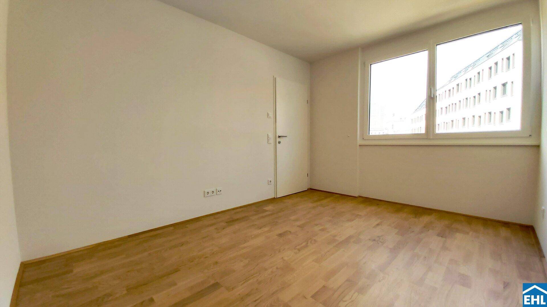 Heller, leerer Raum mit Holzfußboden und einem Fenster, das Tageslicht hereinlässt.