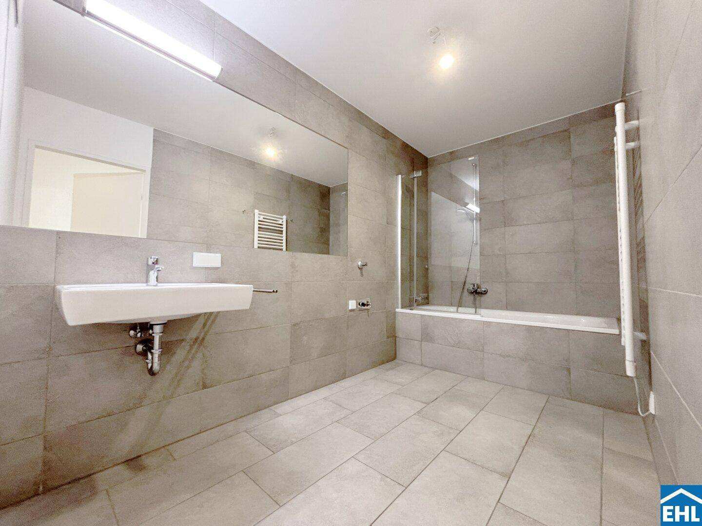 Modernes Badezimmer mit grauen Fliesen, Badewanne, Einzelwaschbecken und beheiztem Handtuchhalter.