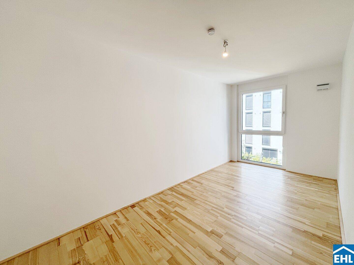Geräumiges, helles Zimmer mit Holzboden und einem großen Fenster, das viel Tageslicht hereinlässt.