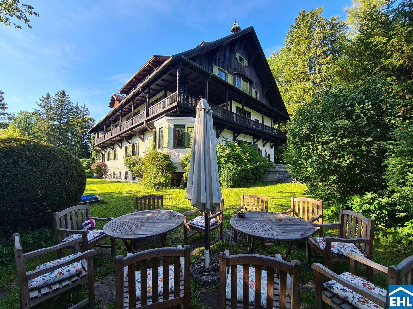 Gemütlicher Sitzbereich im Garten mit Holzmöbeln und Sonnenschirm vor dem historischen Gebäude.