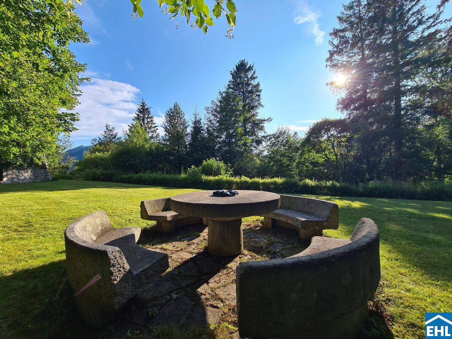 Rustikaler Steinsitzplatz im weitläufigen Garten, umgeben von Bäumen und Natur.