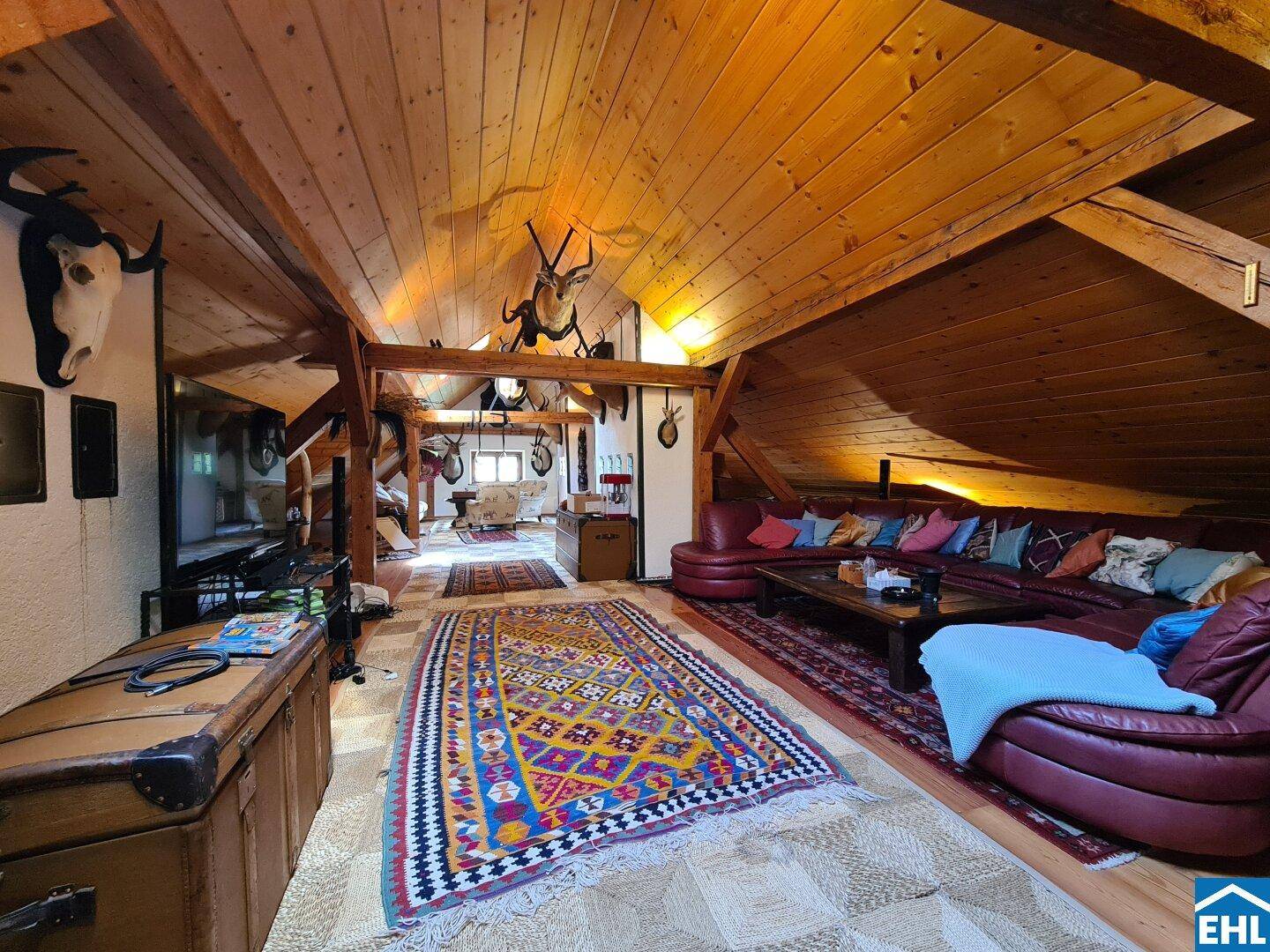 Geräumiges Wohnzimmer im Dachgeschoss mit Holzbalken, rustikalen Teppichen und Jagdtrophäen.
