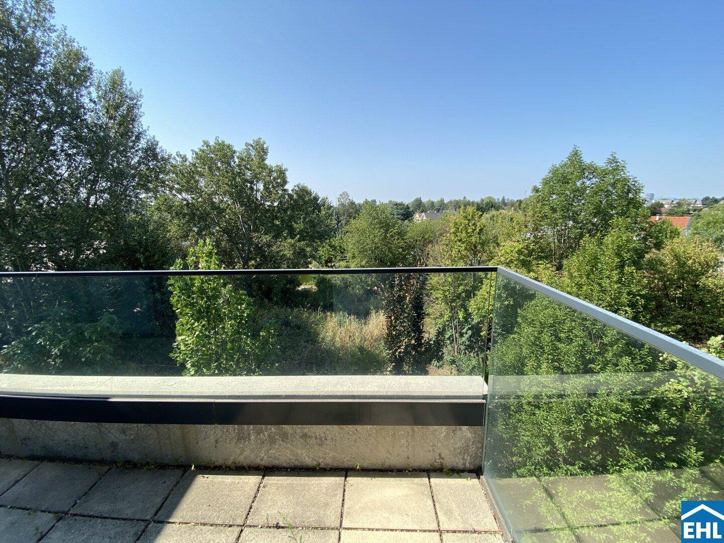 Geräumiger Balkon mit Glasgeländer und weitem Blick ins Grüne unter blauem Himmel.