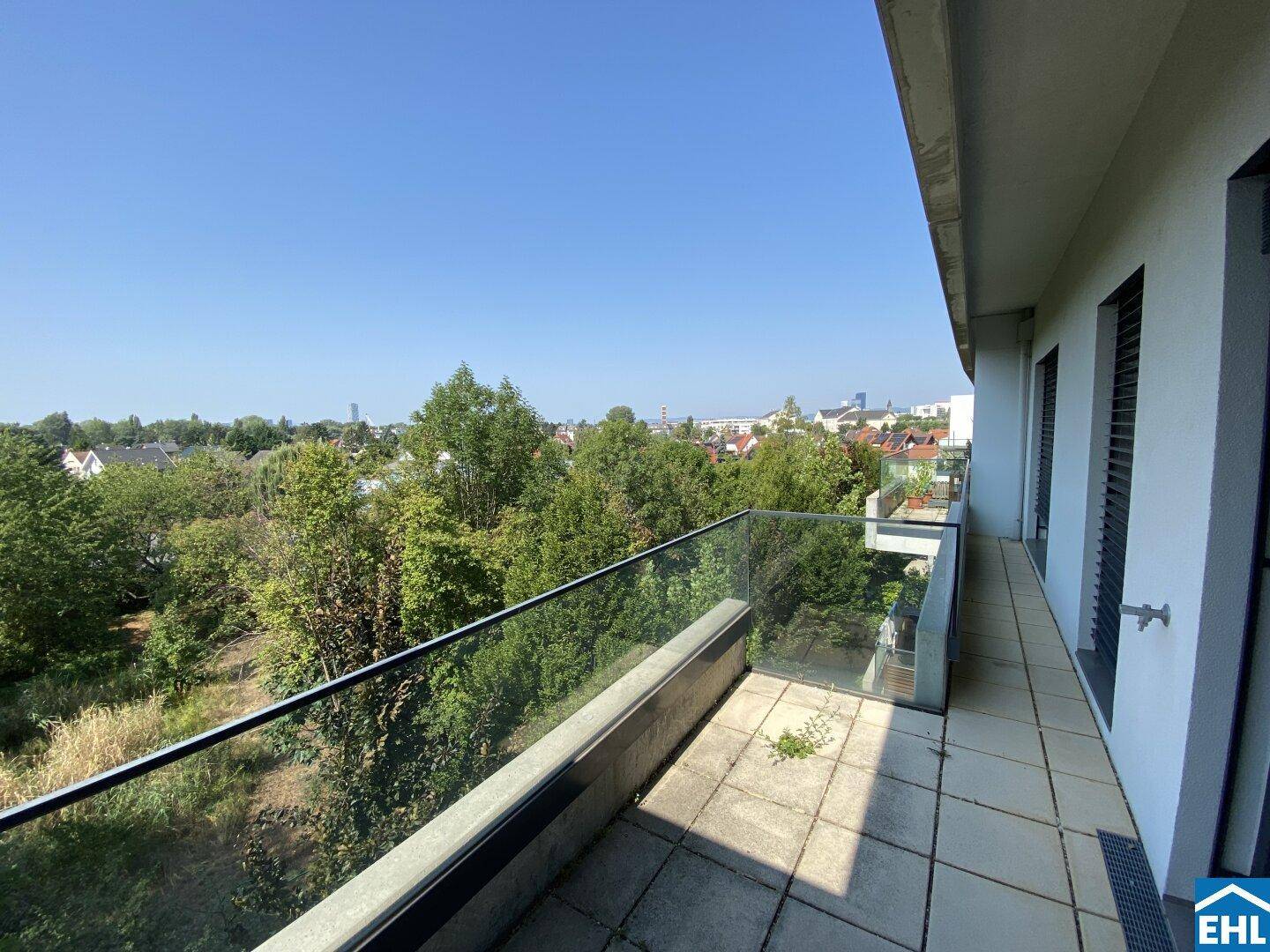 Langer Balkon mit Glasgeländer und freiem Blick auf die grüne Umgebung und die Stadt in der Ferne.