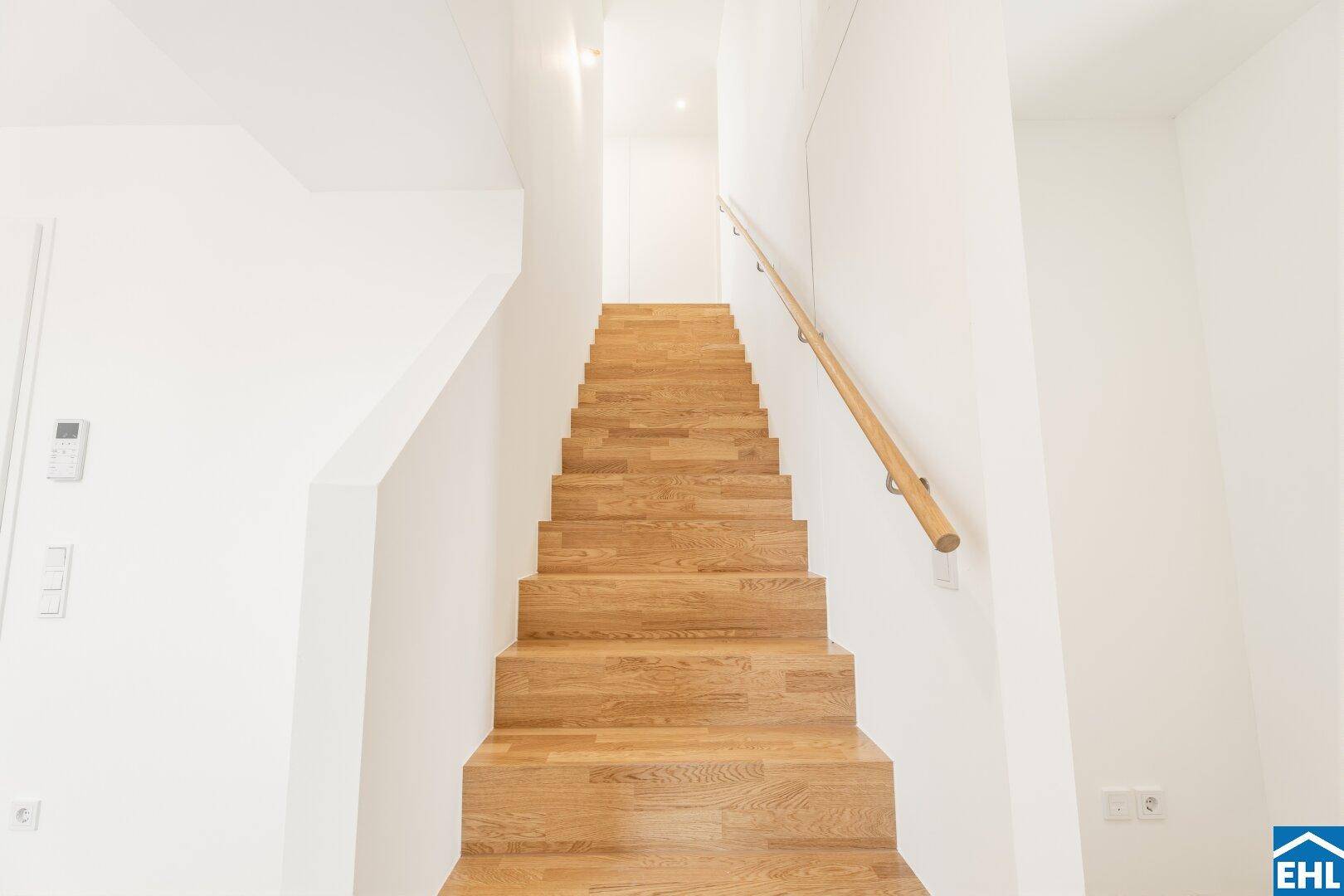 Moderne Holztreppe mit hellem Holzgeländer und weißen Wänden in einem lichtdurchfluteten Treppenhaus.
