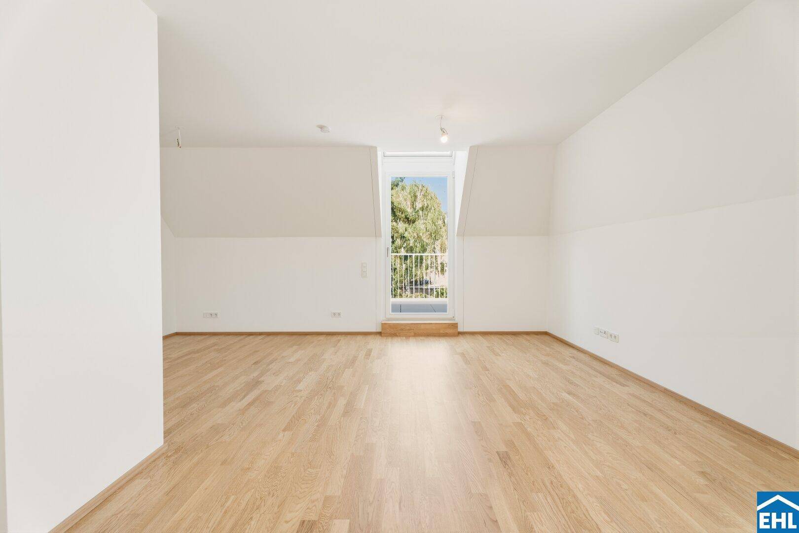 Heller Raum mit Holzboden, schrägen Wänden und einem französischen Balkon, der viel Licht hereinlässt.