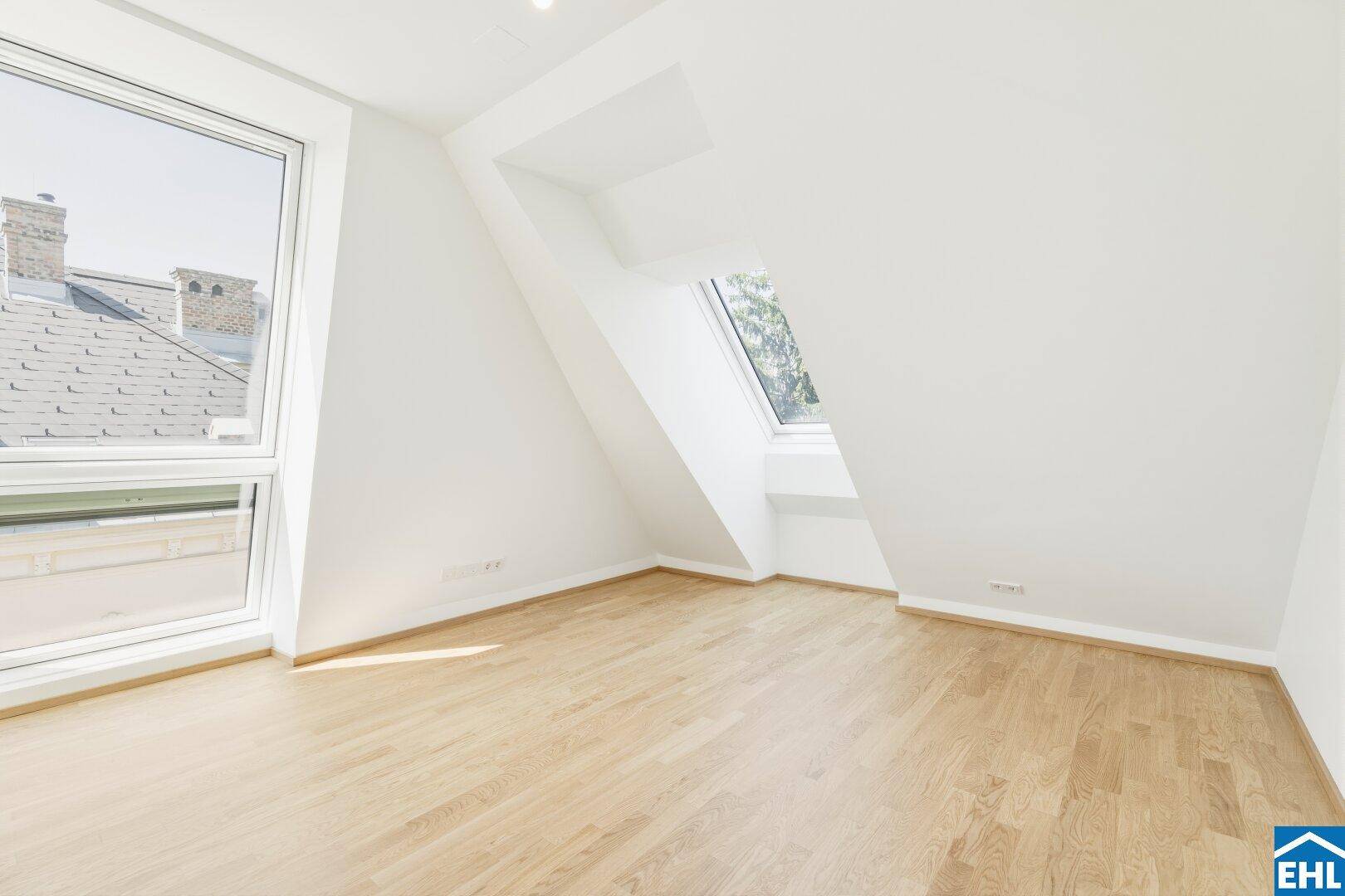 Lichtdurchfluteter Raum im Dachgeschoss mit Holzboden, schrägen Wänden und zwei Fenstern.