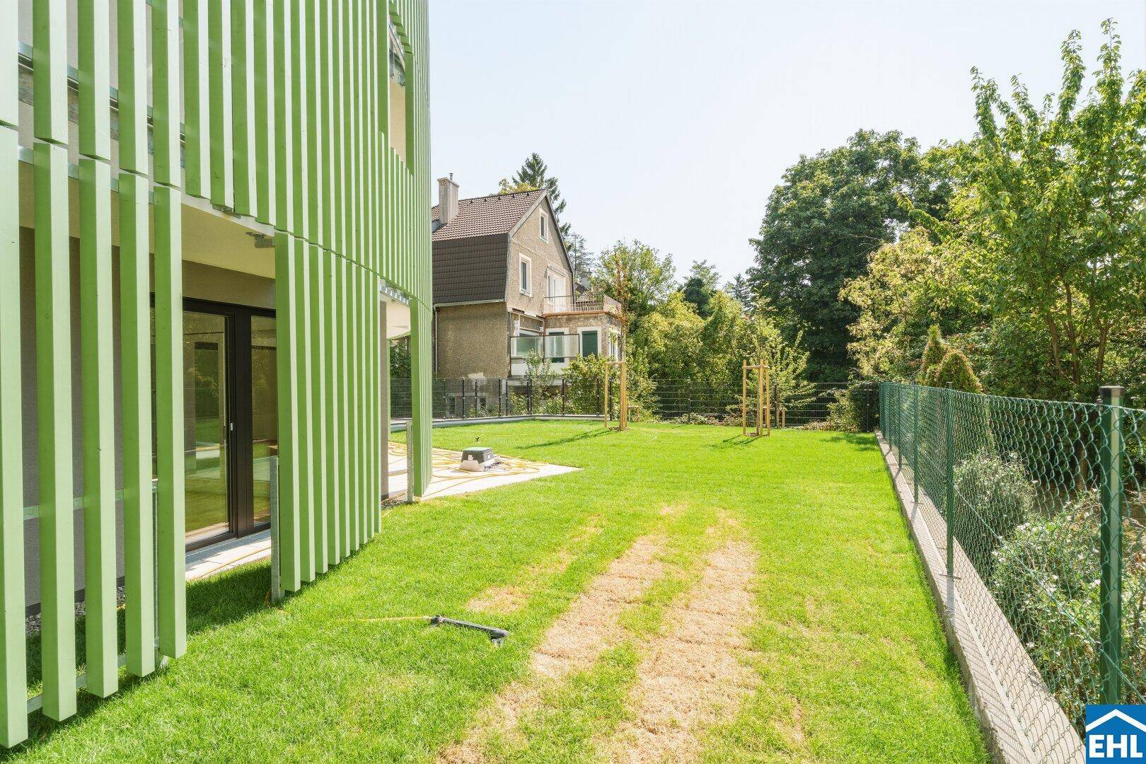 Weitläufiger Gartenbereich neben dem Gebäude mit junger Bepflanzung und Zaun.