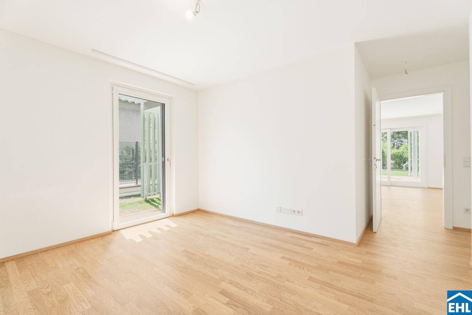 Helles Zimmer mit Holzboden und Tür zum Außenbereich, verbunden mit einem weiteren Raum.