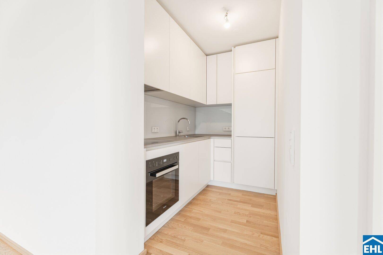 Moderne, kompakte Küche mit weißen Schränken, integriertem Backofen und hellem Parkettboden.