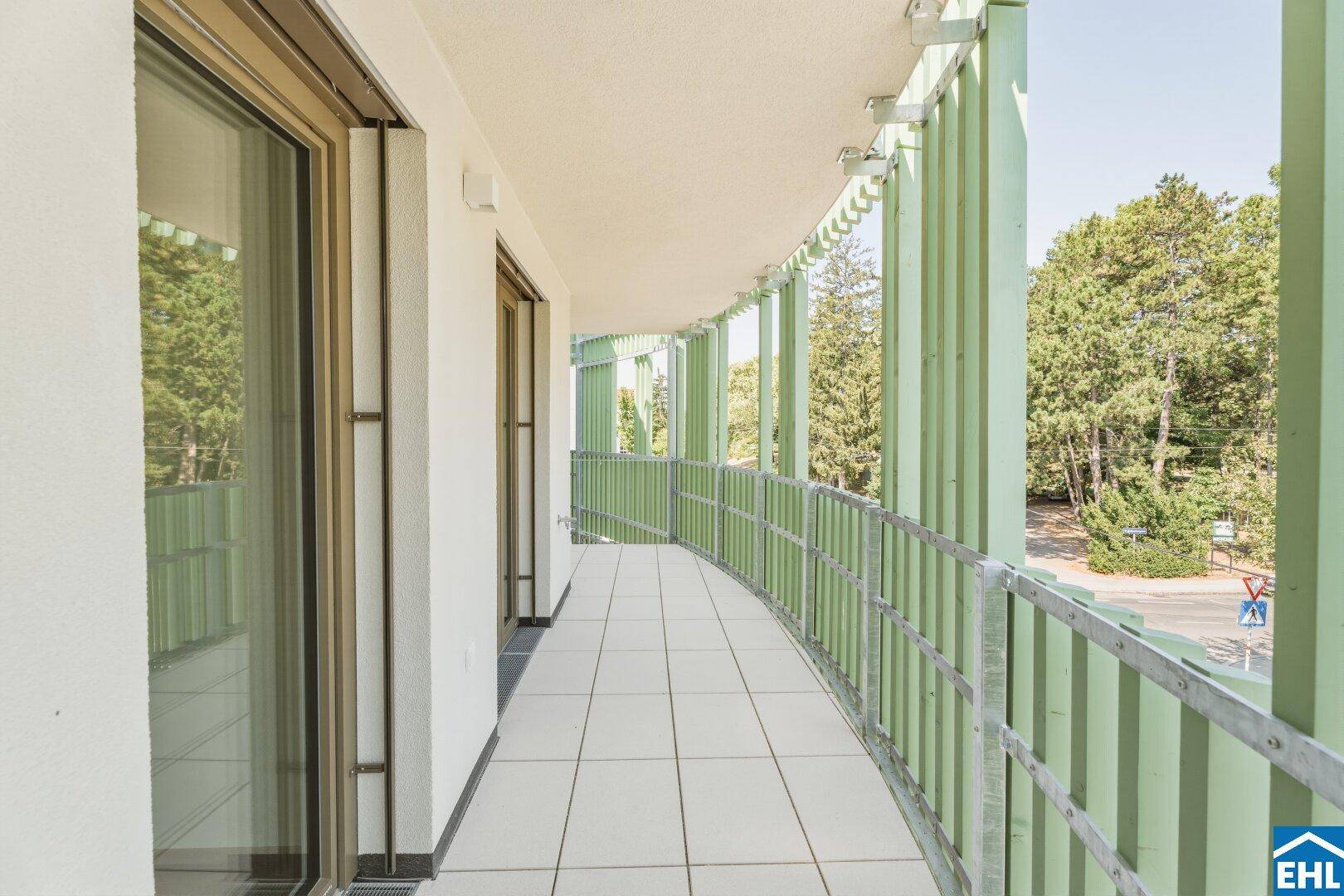 Langer, geschwungener Balkon mit Fliesenboden und grünen Akzenten, umgeben von Bäumen.